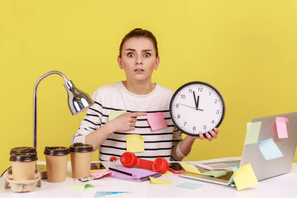 istock Woman worker pointing finger at big wall clock in her hands sitting at workplace, working late hours 1426024381