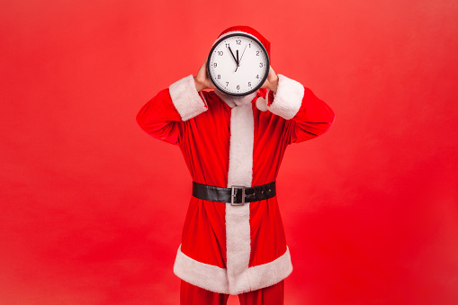 Portrait of unknown man in santa claus costume standing covering face with wall clock, wasting his time, procrastination, organization of working time. Indoor studio shot isolated on red background.