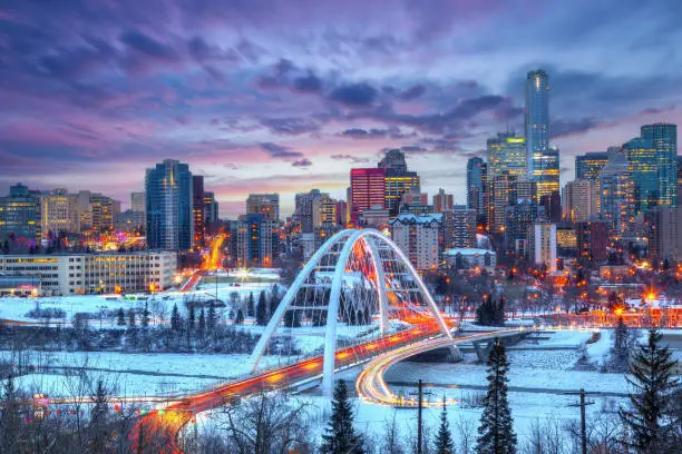 Light trails from rush hour traffic light up Edmonton downtown Winter sunset skyline showing Walterdale Bridge across the frozen, snow-covered Saskatchewan River and surrounding skyscrapers.