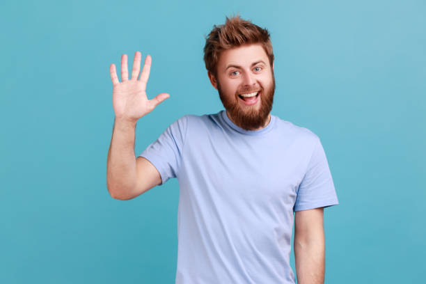 Man in blue T-shirt greeting you rising hand and waving saying hi, glad to see you with toothy smile stock photo