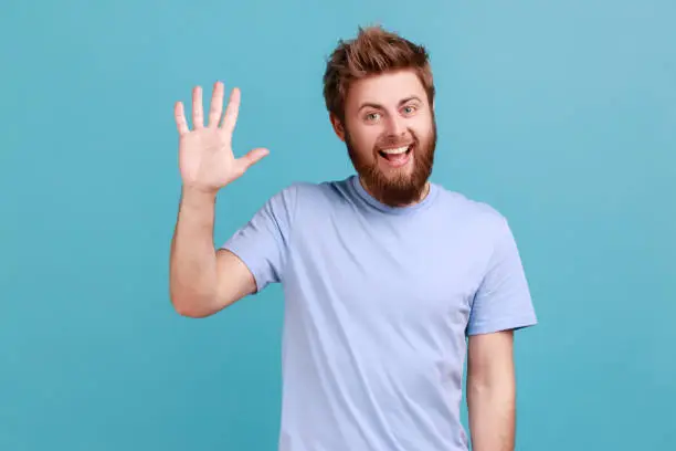 Photo of Man in blue T-shirt greeting you rising hand and waving saying hi, glad to see you with toothy smile