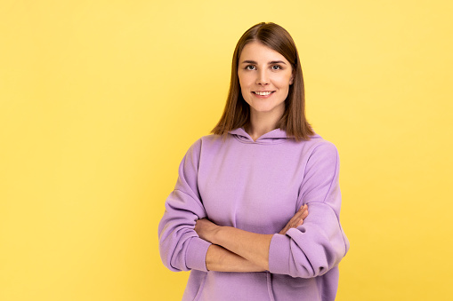 Portrait of happy successful woman standing with crossed hands, looking at camera with charming and joyous toothy smile, wearing purple hoodie. Indoor studio shot isolated on yellow background.