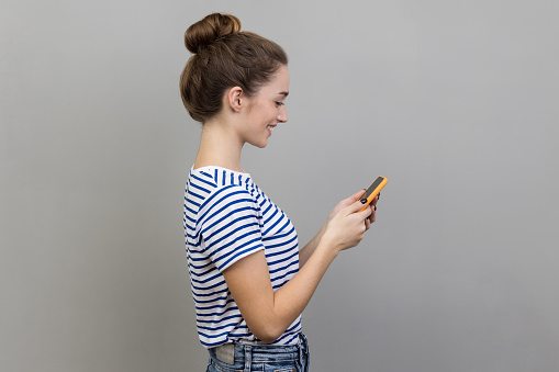 Side view of happy pretty woman wearing striped T-shirt reading message on smartphone and smiling, using mobile device for communication, browsing web. Indoor studio shot isolated on gray background.