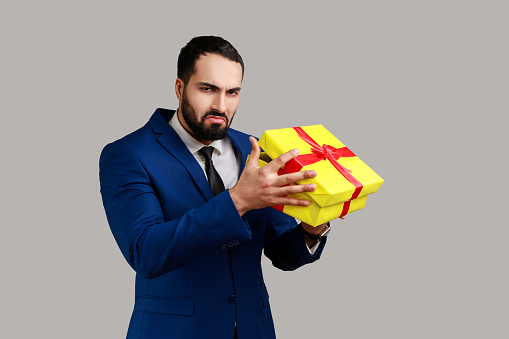 Bearded man opening gift box, looking inside with disappointed sad expression, unwrapping bad present, celebrating holiday, wearing official style suit. Indoor studio shot isolated on gray background.