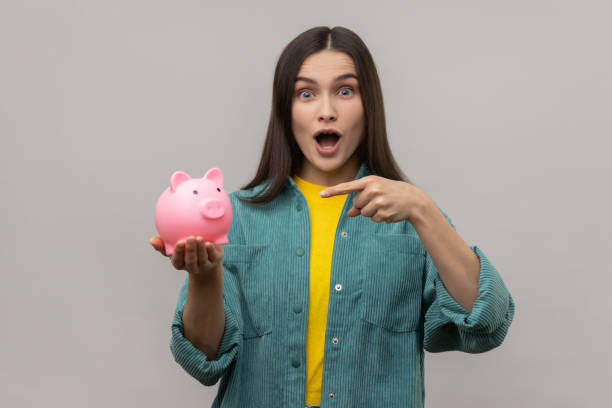 Amazed woman with dark hair standing pointing at piggy bank in her hand, advantageous bank offer. Portrait of amazed woman with dark hair standing pointing at piggy bank in her hand, advantageous bank offer, wearing casual style jacket. Indoor studio shot isolated on gray background. a penny saved stock pictures, royalty-free photos & images