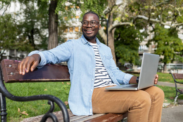 istock Bright male student sitting on park bench with laptop 1426028216