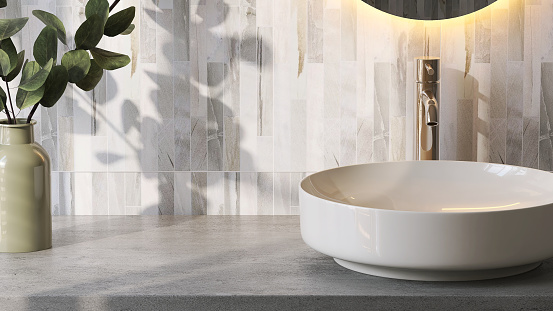 Modern and luxury bathroom vanity counter top with white round ceramic washbasin, gold faucet and frameless mirror with lighting in sunlight from window with vase of twig and granite tiles wall for product display