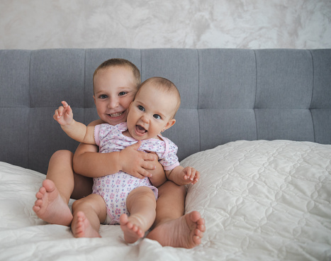 Toddler brother holding his six month old sister sitting on a bed