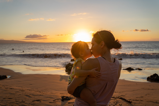A two year old girl is enjoying quality bonding time at the beach while on vacation with her thirty-something year old mom. The Hawaiian mother is holding her daughter and giving her a kiss while the sun sets in front of them.