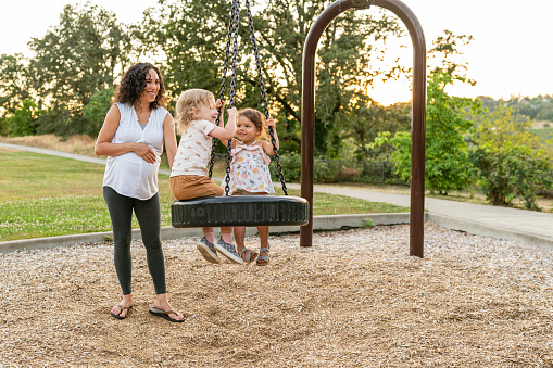 A gorgeous mixed race woman of Pacific Islander descent who is eight months pregnant pushes two happy toddler girls on a tire swing at the park on a warm summer evening.