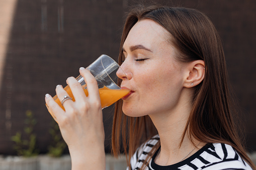 Portrait of beautiful young woman drinking and taking sips of fresh cold orange juice outdoor on terrace closeup side view. Lady with closed eyes enjoying taste of drink, refreshing on summer day.