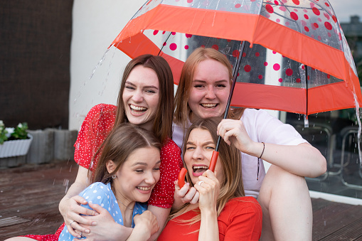 Group of young joyful attractive women sitting on wooden veranda, hiding from rain under umbrella with red dots, enjoying summer weather, having fun. Friendship, relationship, summer, hen-party.