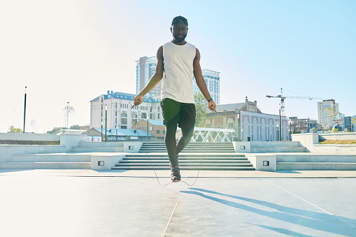 Young active African American man in sportswear jumping over skipping rope while exercising on wide sunlit square in the morning