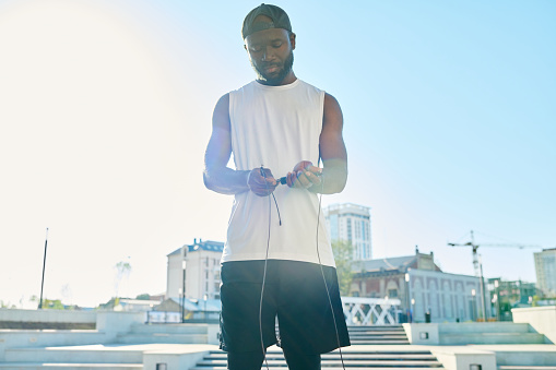 Young serene African American man in sportswear holding skipping rope while standing in front of camera against urban environment
