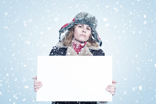Frozen young woman in winter clothes holds an empty white poster in her hands, it is snowing on a blue background