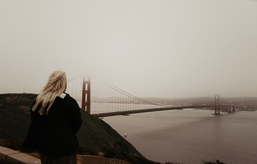 Person looking at the famous Golden Gate Bridge that is barely visible due to fog and cloudy weather. Blonde woman and tourist looking at landscape view of San Francisco Bay, California, USA.