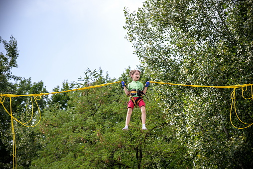 The boy is jumping on a bungee trampoline. A child with insurance and stretchable rubber bands hangs against the sky. The concept of happy childhood and games in the amusement park.