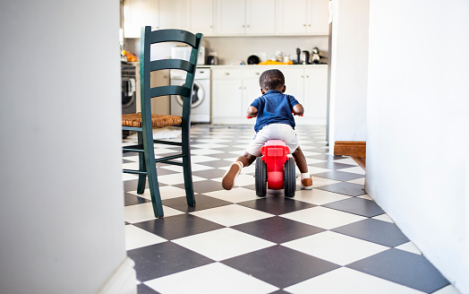 Rear view of a cute little African boy riding his toy tricycle around his kitchen at home