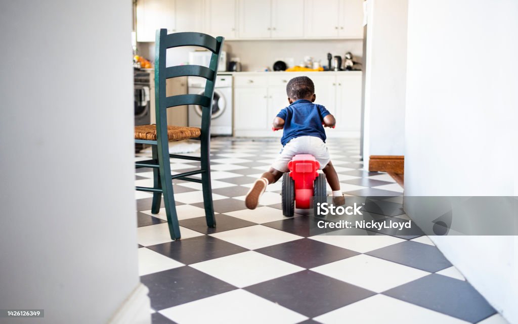 Little boy riding his toy tricycle in a kitchen Rear view of a cute little African boy riding his toy tricycle around his kitchen at home Rear View Stock Photo