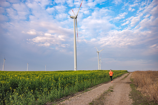 Jogging on dirt road in the agricultural field with wind turbines under the cloudy blue sky