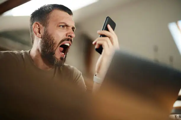 Photo of Angry man yelling while using cell phone while working at home.