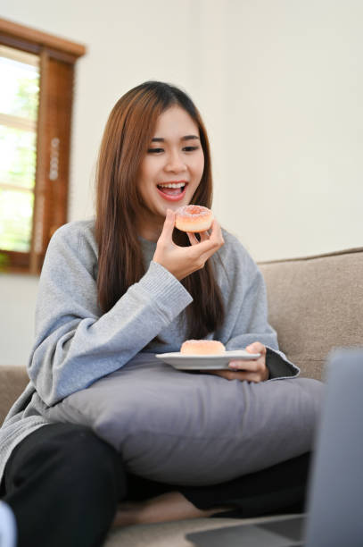 istock Happy Asian female snacking, eating doughnut in her living room while relaxing 1426138788