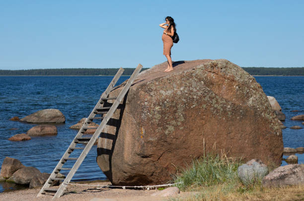 Heavily pregnant brunette woman standing on a big boulder with a ladder on it by the sea, wearing a black backpack and beige tight dress and looking into the distance on Käsmu beach in Estonia stock photo