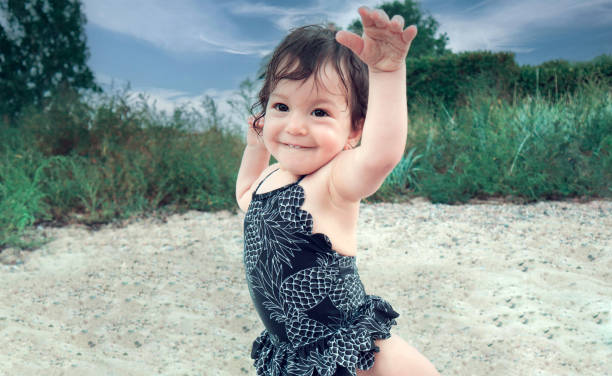 Happy, smiling, adorable brunette baby girl with dark brown eyes on the sandy beach wearing black swimsuit and holding her hands up on a family holiday. stock photo