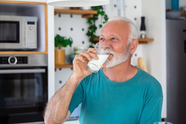starszy mężczyzna pijący szklankę mleka ze szczęśliwą twarzą stojącą i uśmiechniętą. przystojny starszy mężczyzna pijący szklankę świeżego mleka w kuchni - retirement senior adult breakfast active seniors zdjęcia i obrazy z banku zdjęć