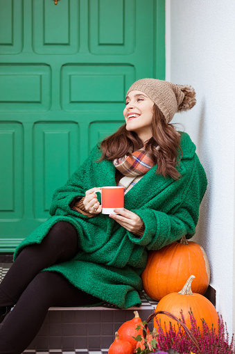 Beautiful woman wearing warm clothes - knit hat, coat ad scarf, sitting in front of house against green door next pumpkins, holding cup of hot tea. Autumn season.