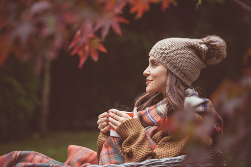 Side view of beautiful woman wearing warm clothes knit hat ad scarf, sitting with cup of tea in autumn garden.