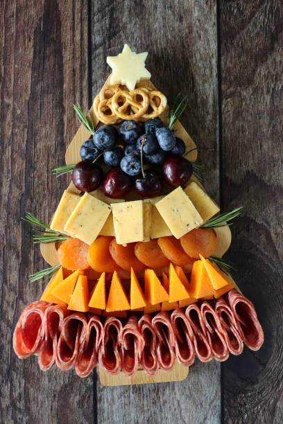 Stock photo showing elevated view of wooden charcuterie board in the shape of a Christmas tree covered with prepared sliced and chopped ingredients including pretzels, blueberries, cherries, cheddar squares, apricots, triangles of Red Leicester cheese, folded salami slices, topped with cheese star. imagen en primer plano de la tabla de charcutería de madera en forma de árbol de navidad cubierta con pretzels, arándanos, cerezas, cuadrados de cheddar, albaricoques, triángulos de queso red leicester, rodajas de salami dobladas, cubiertas con estrell