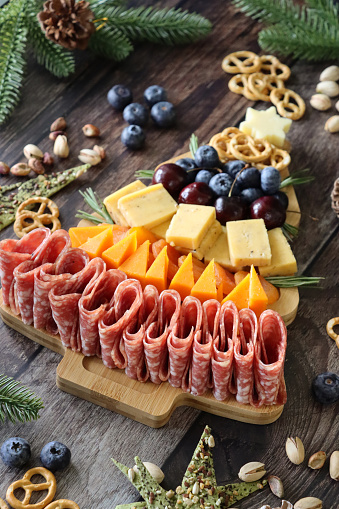 Stock photo showing close-up view of Christmas tree, wooden charcuterie board covered with prepared sliced and chopped ingredients including pretzels, blueberries, cherries, cheddar squares, apricots, triangles of Red Leicester cheese, folded salami slices, topped with cheese star and surrounded by spruce tree needles, pistachios, pine cones and green triangle crackers.