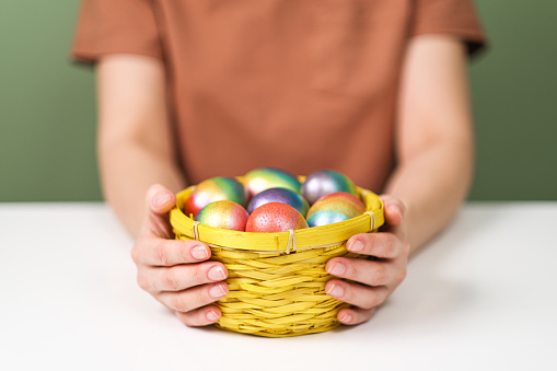 A young beautiful woman in a brown t-shirt paints Easter eggs in different colors. Preparing Easter Eve at home. Green wall background.