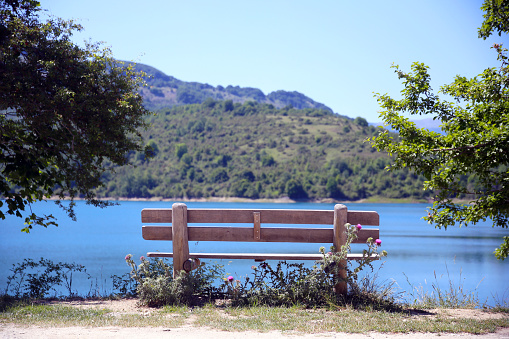 Behind the wooden bench, among the trees, overlooking the blue waters of the lake and the tree-lined mountains, Lago della Montagna Spaccata, Alfedena, Abruzzo, Italy