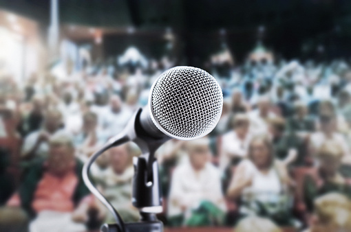 Vocal microphone awaiting public speaker, singer or actor in a crowded theater, with blurred audience waiting for the show
