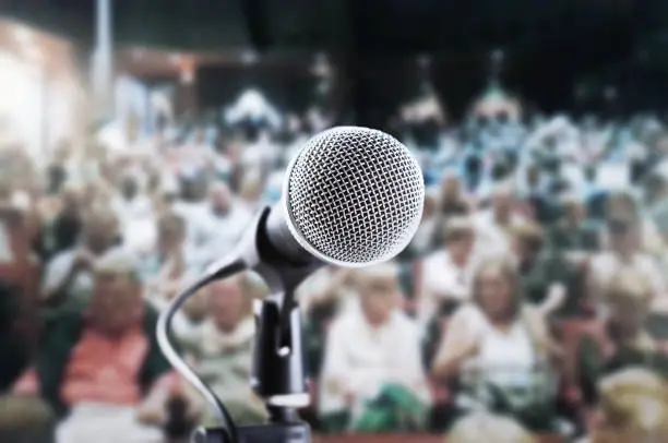 Photo of Vocal microphone awaiting public speaker, singer or actor in a crowded theater, with blurred audience waiting for the show