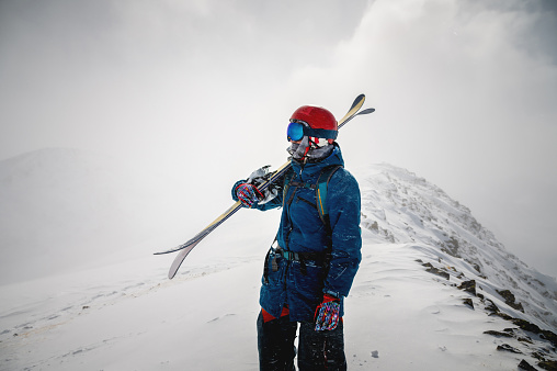 The skier holds a pair of skis and looks at the snow-capped mountains. A guy with skis on his shoulder stands in the mountains covered with snow. Portrait of a man with winter gear.