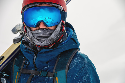 Equipped skier holding skis on his shoulder and looking directly at the camera, portrait. Man with winter equipment in the mountains at a ski resort.