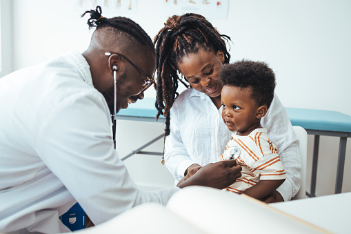 A happy toddler boy sits in his mother' lap at the pediatrician. He plays patacake with a doctor. Doctor giving prescription to african american mother with baby son at clinic. Medicine, healthcare and pediatry concept.
