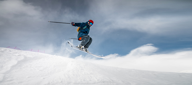 A sportsman skier in ski equipment jumps down a steep snowy slope of a mountain against the backdrop of a blue sky and snow-capped mountains. Winter risky sports, courage and speed concept.