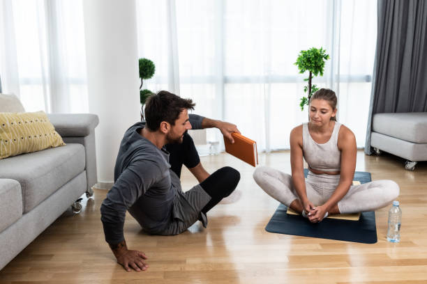 giovane istruttore di fitness e yoga che lavora a casa tua. uomo terapista allenatore istruito sportivo che aiuta la giovane donna a riprendersi nel suo appartamento e a casa in allenamento e allenamento per una vita sana. - yoga men male gymnastics foto e immagini stock