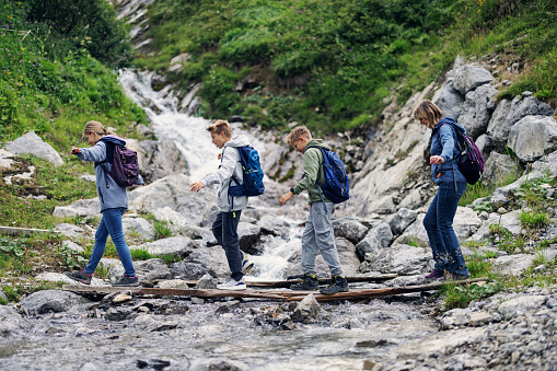 Mother and three teenage kids are hiking in the river valley in the Alps - Tyrol, Austria. They are walking on the path and crossing a stream over a small bridge.\nCanon R5
