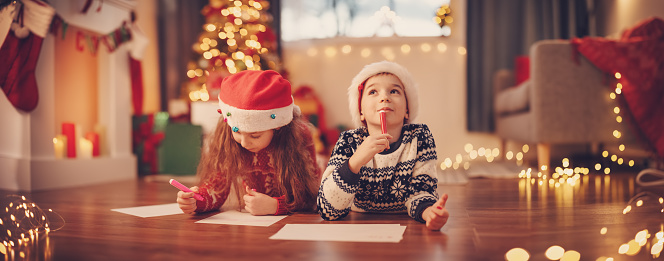 Children lying on the floor indoors and writing letters for Santa Claus. Concept of the Christmas eve.