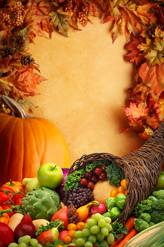 A Thanksgiving cornucopia of fruits and vegetables and a holiday pumpkin in front of a textured wall framed by autumn colored leaves.