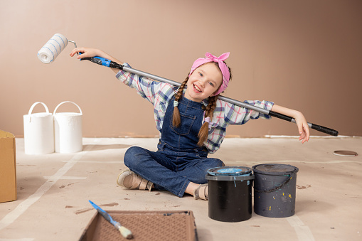 A cute little girl plays with a paint roller while renovating a room. The sweet child spreads hands puts the stick behind them and smiles at the camera.