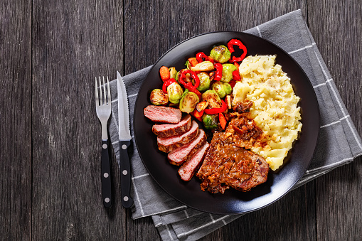 medium rare beef steak with brown gravy and mashed potato, roasted brussels sprouts, pepper and carrots on black plate on dark wooden table with cutlery, flat lay