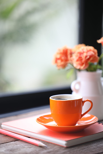 Orange roses and small coffee cup and noteebook pencil on wooden in front of windows