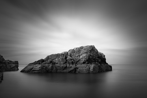 Small rocky island in Howth, Dublin, captured in long exposer, blurred water and clouds, converted to fine art black and white photo, Ireland
