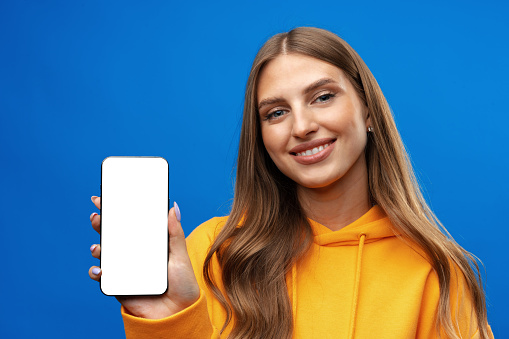 Young woman showing big blank cellphone screen on blue background in studio, copy space